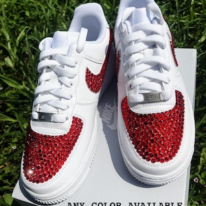 May include: White sneakers with red crystal embellishments on the toe and Nike swoosh. The shoes have white laces and a silver "AF1" tag. The text "ANY COLOR AVAILABLE" is printed at the bottom of the image.