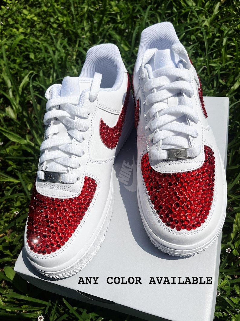 May include: White sneakers with red crystal embellishments on the toe and Nike swoosh. The shoes have white laces and a silver "AF1" tag. The text "ANY COLOR AVAILABLE" is printed at the bottom of the image.
