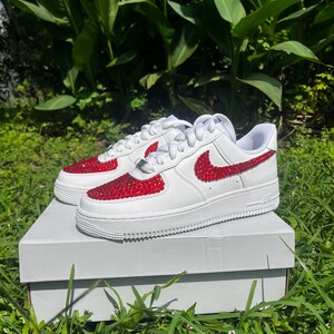 May include: White sneakers with red rhinestone accents on the toe and Nike swoosh. The shoes are on a white box, set against a backdrop of green foliage and grass.