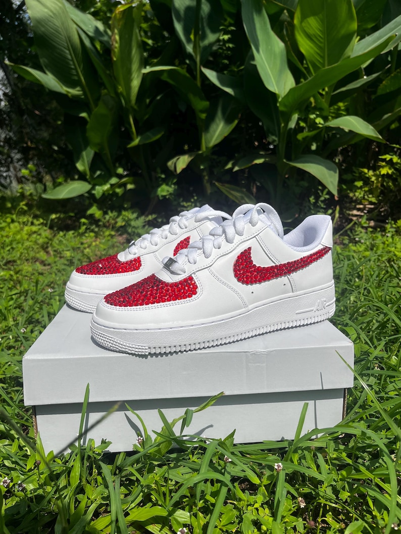 May include: White sneakers with red rhinestone accents on the toe and Nike swoosh. The shoes are on a white box, set against a backdrop of green foliage and grass.
