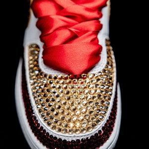 May include: White sneakers with red and gold rhinestones. The sneakers have a red satin ribbon tied around them.