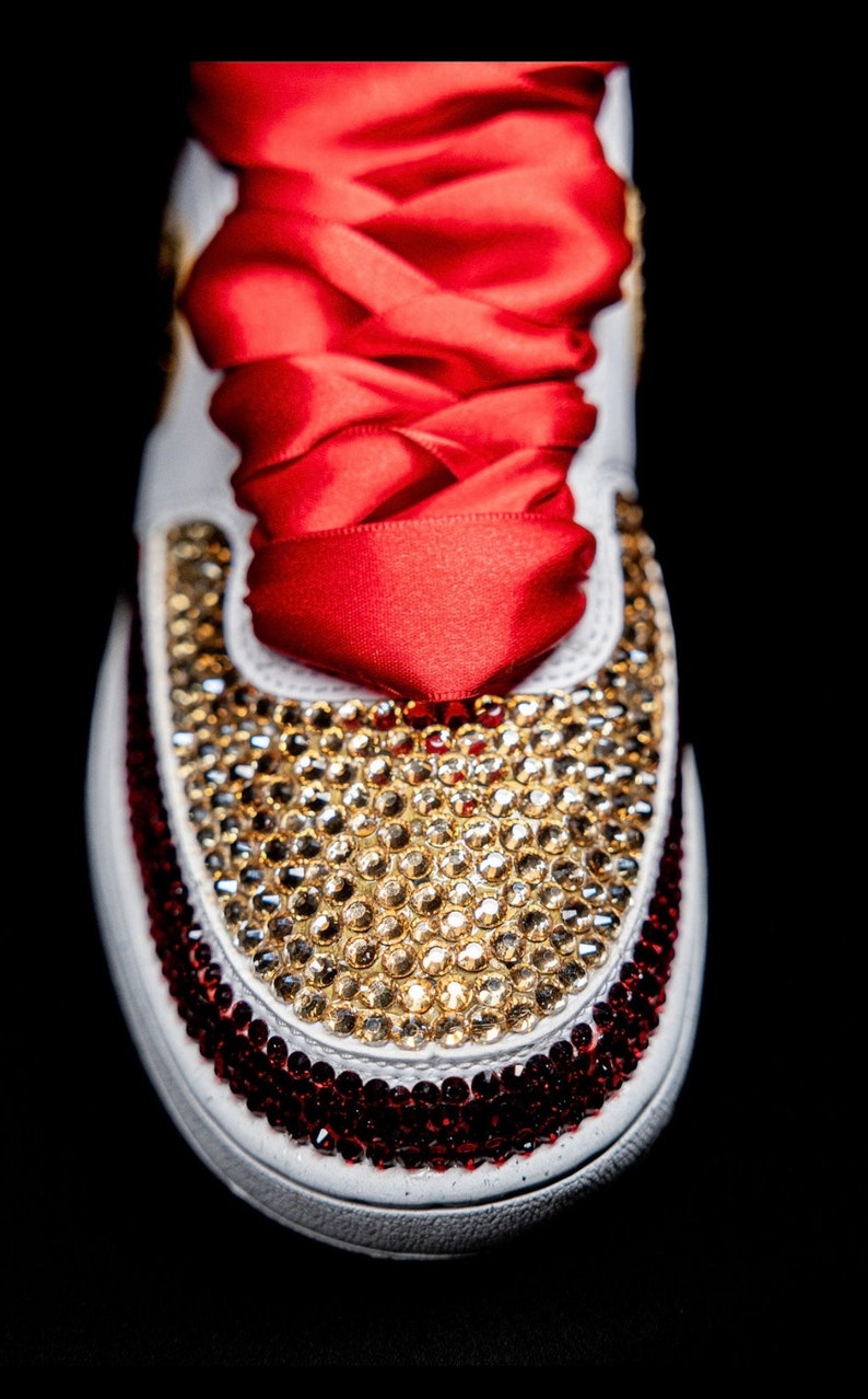 May include: White sneakers with red and gold rhinestones. The sneakers have a red satin ribbon tied around them.