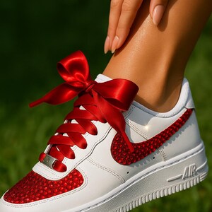 May include: White leather sneaker with red jewel accents on the toe and Nike swoosh. The laces are replaced with a red satin ribbon tied in a bow. The shoe has a white sole and the word "AIR" is visible.