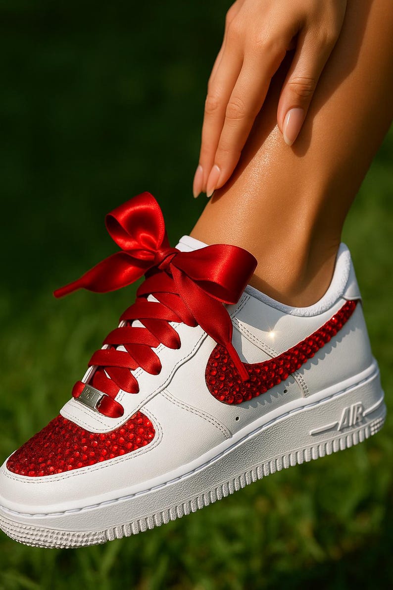 May include: White leather sneaker with red jewel accents on the toe and Nike swoosh. The laces are replaced with a red satin ribbon tied in a bow. The shoe has a white sole and the word "AIR" is visible.