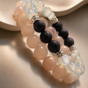 Può includere: Tre braccialetti di perline. Un braccialetto presenta perline rosa chiaro, un altro ha perline di lava nere e il terzo ha perline bianche e iridescenti. Distanziatori color argento sono tra le perline. I braccialetti sono impilati.