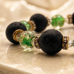 May include: Close-up of a bracelet featuring black lava rock beads, clear beads with green and gold inclusions, and gold-toned spacers. The bracelet is displayed on a light-colored surface.