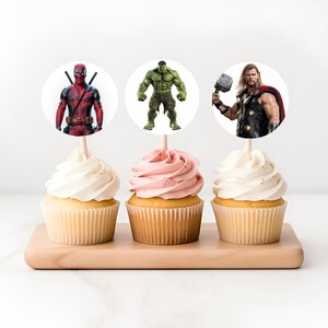 May include: Three cupcakes decorated with superhero themed toppers. The cupcakes are topped with white and pink frosting. The toppers feature Deadpool, Hulk, and Thor.