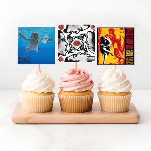 May include: Three cupcakes with frosting, each topped with a different album cover. The cupcake on the left has a blue and white album cover with the text "Nirvana" and "Nevermind". The middle cupcake has a black and white album cover with a red rose and the text "Red Hot Chili Peppers". The cupcake on the right has a yellow and red album cover with the text "Guns N' Roses" and "Use Your Illusion I".
