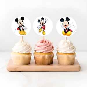 Puede incluir: Tres cupcakes con glaseado blanco y rosa cubiertos con adornos redondos de Mickey Mouse. Los adornos muestran a Mickey Mouse en diferentes poses.