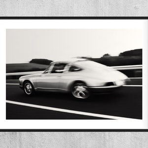 Puede incluir: Una fotografía en blanco y negro de un coche vintage plateado conduciendo por una carretera. El coche está en movimiento, creando un efecto de desenfoque. El fondo está desenfocado, mostrando un campo y árboles.