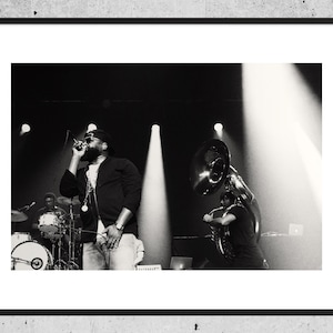 Peut inclure: Une photographie en noir et blanc d'un musicien chantant dans un microphone sur scène. Le musicien porte une veste noire, un t-shirt blanc et une casquette noire. Un joueur de tuba se tient à droite du chanteur. La scène est éclairée par des projecteurs.