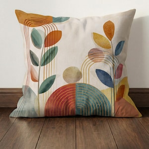 May include: Square decorative pillow with an abstract botanical design. The pillow features colorful shapes and lines in shades of orange, blue, green, and yellow on a cream background. The design includes leaf shapes and arched lines.
