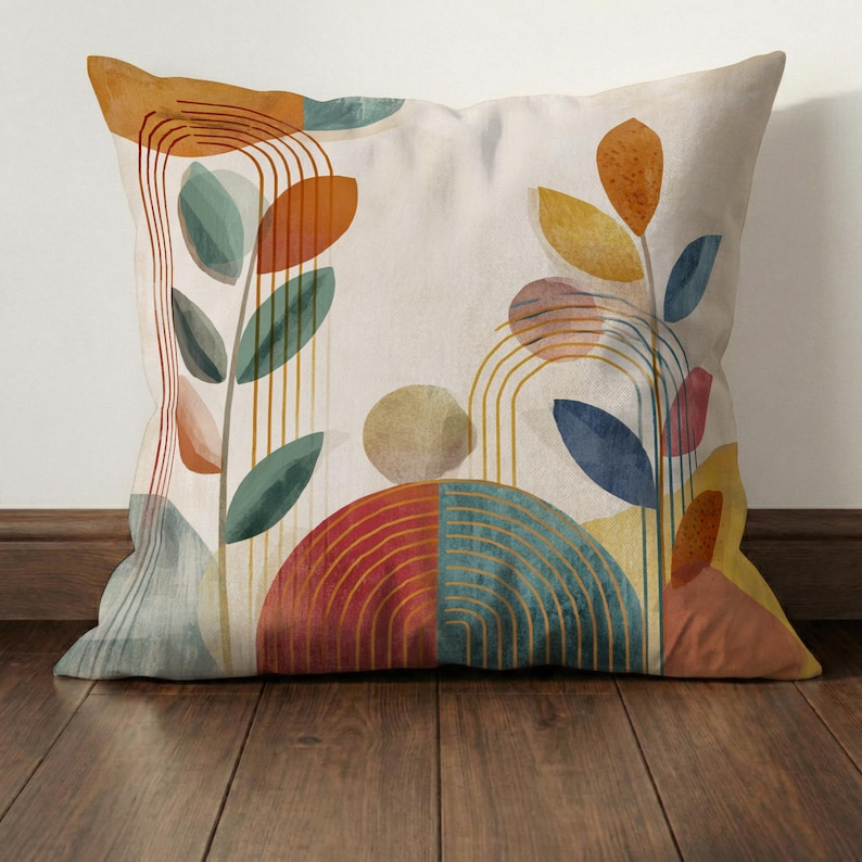 May include: Square decorative pillow with an abstract botanical design. The pillow features colorful shapes and lines in shades of orange, blue, green, and yellow on a cream background. The design includes leaf shapes and arched lines.