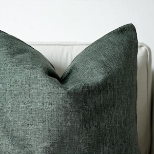 Könnte beinhalten: Nahaufnahme eines dunkelgrünen Zierkissens mit strukturiertem Stoff. Das Kissen hat eine quadratische Form und wirkt weich und einladend. Das Kissen liegt auf einer weißen Oberfläche.