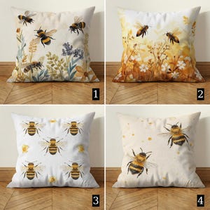 May include: Four square cushions with bee-themed designs. Each cushion features a different watercolour illustration of bees and floral elements. The cushions are on a light-coloured surface.