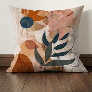 May include: Square decorative pillow with an abstract design. The pillow features a beige background with orange, brown, and teal shapes, including a dark blue circle and a stylized leaf. The design has a modern, artistic aesthetic.