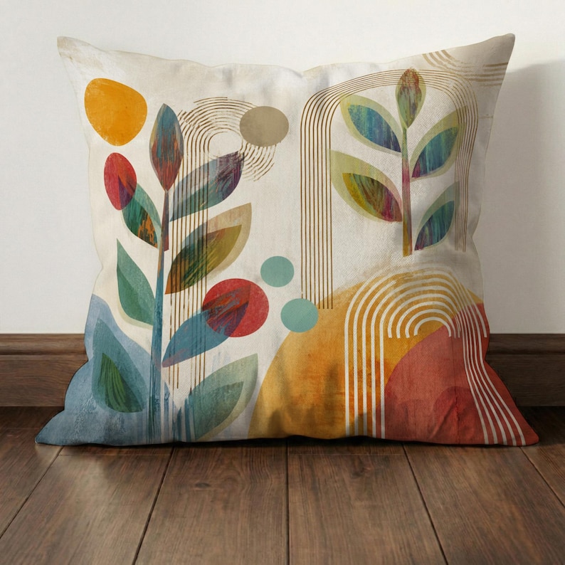 May include: Square decorative pillow with an abstract botanical design. The pillow features colorful leaves, circles, and arched lines in shades of orange, red, blue, and green against a cream background. The pillow is placed on a wooden floor.