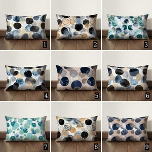 Puede incluir: Nueve cojines rectangulares con diseños abstractos. Cada cojín presenta un patrón diferente de círculos superpuestos y lavados de estilo acuarela en varios colores, incluyendo azul, negro y beige. Los cojines se exhiben sobre un suelo de madera.