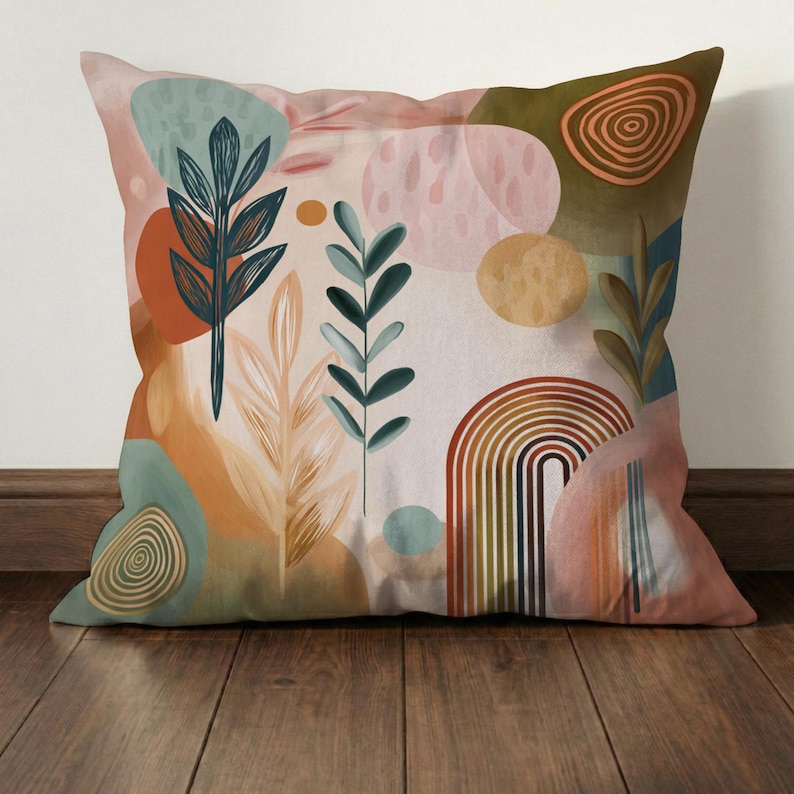 May include: Decorative square pillow with an abstract botanical design. The pillow features a cream background with colorful shapes, including a rainbow, circles, and leaf-like illustrations in shades of orange, green, and pink. The pillow is placed on a wooden floor.