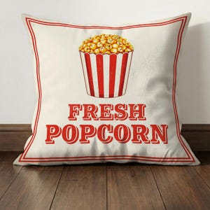 Może przedstawiać: Kwadratowa poduszka z kremowym tłem i czerwoną obw&oacute;dką. Poduszka przedstawia ilustrację w stylu vintage wiadra z popcornem wypełnionego popcornem oraz czerwony napis "FRESH POPCORN".