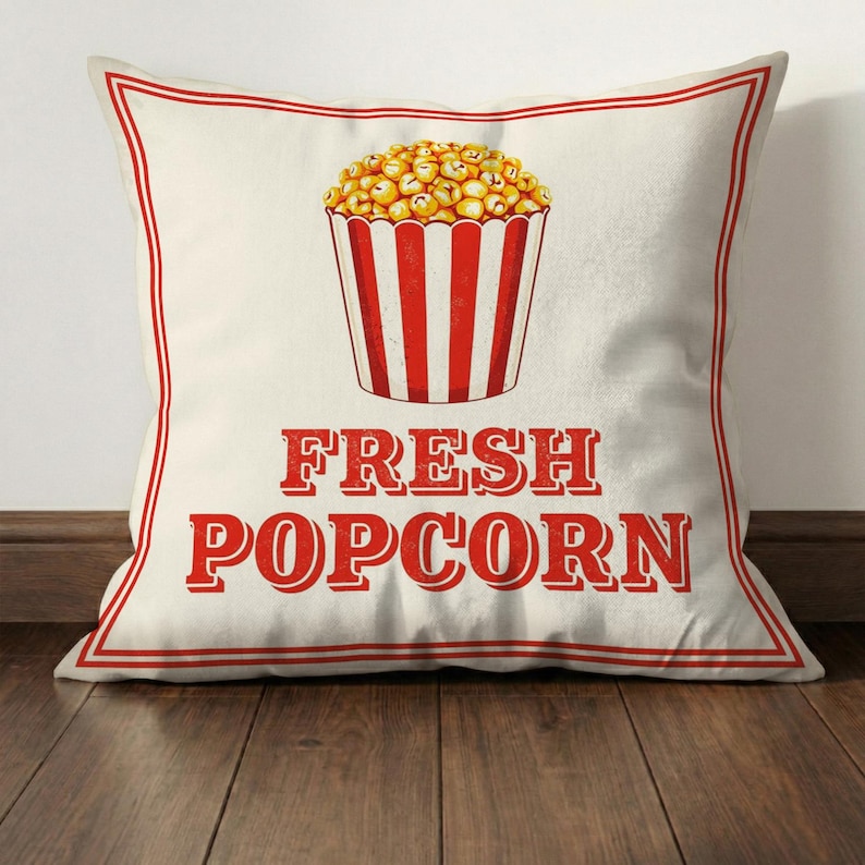 Może przedstawiać: Kwadratowa poduszka z kremowym tłem i czerwoną obw&oacute;dką. Poduszka przedstawia ilustrację w stylu vintage wiadra z popcornem wypełnionego popcornem oraz czerwony napis "FRESH POPCORN".