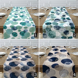 Op de afbeelding: Vier tafellopers met abstracte aquarelontwerpen. Elke loper heeft een ander patroon van overlappende cirkels in tinten blauw, groen en neutrale tinten. De lopers worden gepresenteerd op een houten tafel met borden, glazen en bestek.