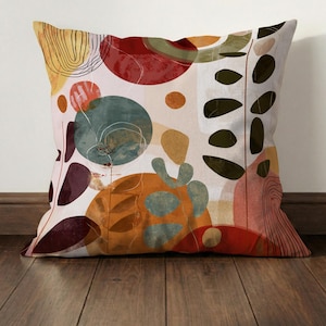May include: Square decorative pillow with an abstract design. The pillow features a cream background with colorful shapes in red, orange, teal, and olive green. Black leaf-like shapes and thin black lines add detail to the design.