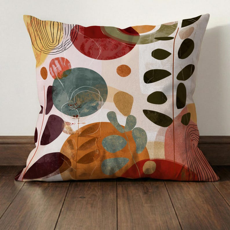 May include: Square decorative pillow with an abstract design. The pillow features a cream background with colorful shapes in red, orange, teal, and olive green. Black leaf-like shapes and thin black lines add detail to the design.