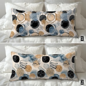 May include: Rectangular decorative pillow with a cream background and a pattern of black, blue, and tan circles and scribbles. The pillow is placed on a white bed with white pillows.