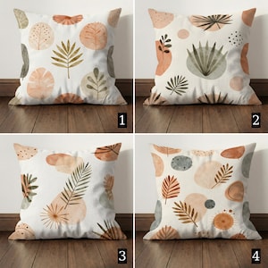 Op de afbeelding: Vier vierkante decoratieve sierkussens met botanische ontwerpen. Elk kussen heeft een witte achtergrond met abstracte vormen en bladpatronen in tinten bruin, perzik en grijs. De kussens zijn gerangschikt op een houten vloer.