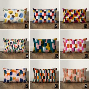 Könnte beinhalten: Neun rechteckige Kissen mit abstrakten Designs. Die Kissen weisen verschiedene Farbpaletten auf, darunter Blau, Gelb, Orange, Rosa und Grün, mit Pinselstrich- und geometrischen Mustern. Jedes Kissen wird auf einer Holzoberfläche präsentiert.