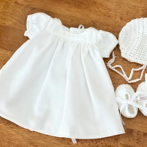 Puede incluir: Un vestido blanco de bebé con mangas cortas y un gorro, zapatos y una cinta de ganchillo.