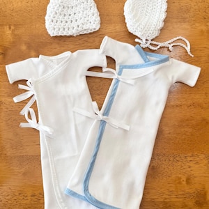 以下が含まれることがあります： 新生児用のベビー服セット。白いラップガウン2着、かぎ針編みのビーニー、ボンネットが含まれています。ガウンの1つは青いトリムが付いています。柔らかい白い生地で作られており、白いリボンで結ばれています。