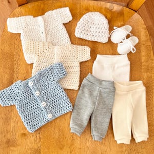 May include: A selection of handmade baby clothes, including crocheted cardigans in white and light blue, a white crocheted hat, white booties, and grey and white trousers. The items are arranged on a wooden surface.