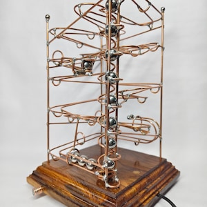 Pode incluir: Um brinquedo de montanha-russa de mármore de cobre e aço com uma base de madeira. A estrutura apresenta uma série de laços, espirais e pistas projetadas para que as bolinhas viajem. A base é de madeira escura e polida.