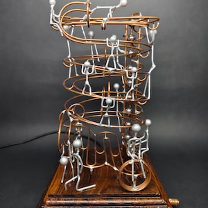 May include: A kinetic sculpture featuring copper wire tracks forming a spiral marble run mounted on a dark wood base. Silver-colored stick figures are positioned along the tracks, with several small silver metal spheres resting on the wire path.