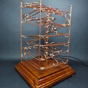 May include: A copper and silver marble run sculpture with a wooden base. The sculpture features a series of loops and tracks for marbles to travel through. The wooden base is square and stained a dark brown color.
