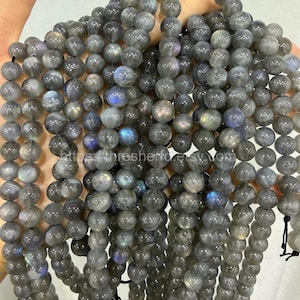 May include: A collection of grey labradorite gemstone beads. The beads are spherical and polished, displaying flashes of blue and iridescent hues. The beads are strung on black thread. Text on the image reads: https://thresherstone.etsy.com