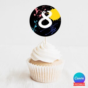 Op de afbeelding: Een zwart-witte cupcake topper met een wit nummer 8 en kleurrijke verfspatten. De topper zit op een witte cupcake met witte frosting.