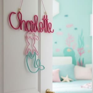 May include: A wall hanging decoration featuring the name Charlotte written in dark pink cursive rope, suspended by a thin string with two wooden beads. Below the name is a wire-framed mermaid silhouette with a light pink torso and teal tail.
