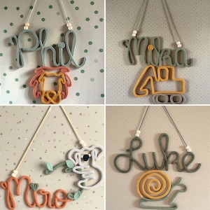 May include: Four wall-hanging name signs crafted from knitted yarn cord. Each sign features a name in cursive script paired with a shape: a dog, an excavator, a koala, and a snail. The designs include wooden beads and hanging strings.