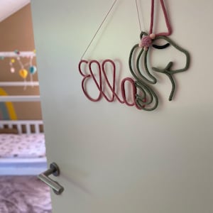 May include: A wall hanging made of rope shaped into the name Ella and a unicorn head. The name is in dusty rose, while the unicorn head is in sage green with a dusty rose horn and a small pom-pom detail. It hangs from a thin string.