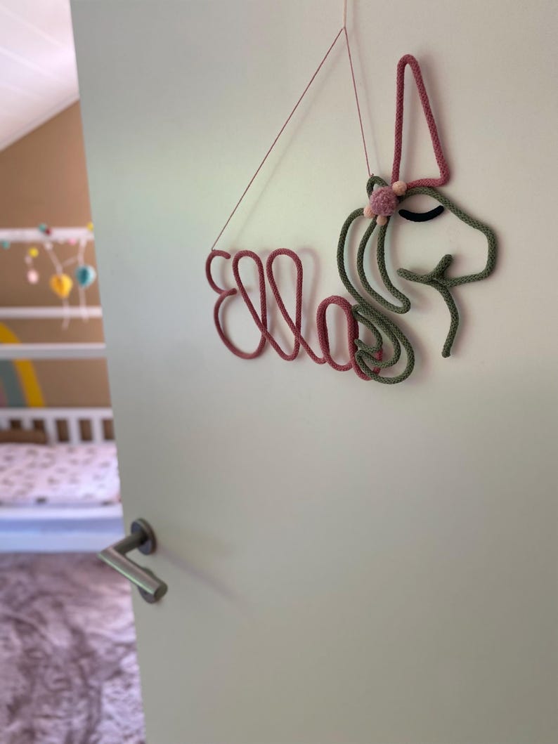 May include: A wall hanging made of rope shaped into the name Ella and a unicorn head. The name is in dusty rose, while the unicorn head is in sage green with a dusty rose horn and a small pom-pom detail. It hangs from a thin string.
