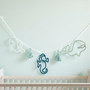 Op de afbeelding: Een nautische slinger met walvis- en zeepaardvormen in wit en blauwgroen, geregen aan een wit koord. De slinger bevat blauwgroene kwastjes en hangt tegen een lichtblauwe muur, boven een wieg.