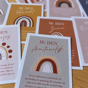 May include: A set of affirmation cards featuring a rainbow design and the text "IK BEN" followed by a positive affirmation in Dutch. The cards are printed on white cardstock with a brown background.