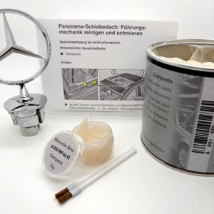 May include: A Mercedes-Benz hood emblem, a 10g container of lubrication paste labeled A 001 989 46 51, a larger metal can of lubrication paste, two small brushes, and an instruction sheet titled Panorama-Schiebedach: Führungsmechanik reinigen und schmieren.