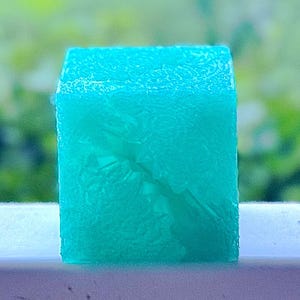 May include: A translucent, bright turquoise cube with a textured, crystalline surface. The object features a geometric shape with sharp edges and a semi-transparent appearance that allows light to pass through its solid, block-like structure.
