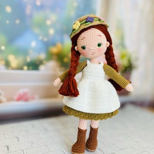 Könnte beinhalten: Handgemachte Häkelpuppe mit roten Zöpfen, grünen Augen und einem Blumenhut. Die Puppe trägt ein weißes Kleid, ein grünes Oberteil mit langen Ärmeln und braune Stiefel. Die Puppe steht auf einer weißen Ziegeloberfläche.
