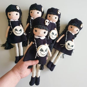 Könnte beinhalten: Fünf gehäkelte Puppen, die schwarze Kleider mit weißen Verzierungen tragen und weiße gehäkelte Handtaschen mit einem schwarz-weißen Totenkopfdesign tragen. Die Puppen haben schwarzes Haar und schwarze Augen.