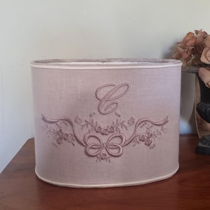 K&ouml;nnte beinhalten: Helllila ovale Lampenschirm mit einem gestickten Design. Das Design umfasst einen gro&szlig;en Buchstaben "C", B&auml;nder und florale Elemente. Der Lampenschirm ist etwa 25 cm hoch.